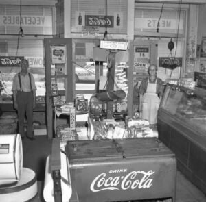 Black and white photo of a vintage store with two people, coca-cola cooler